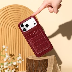 May include: A burgundy phone case with a crocodile texture and a card pocket. The case is held up by a hand, with the text "Ninetydegree" in cursive script at the bottom. The case is on a wooden stump.