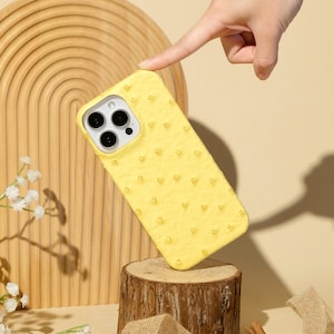 May include: A bright yellow phone case with a textured surface, featuring a camera cutout. The case is displayed on a wooden stand against a neutral background. The text "Ninetydegree" is visible at the bottom.