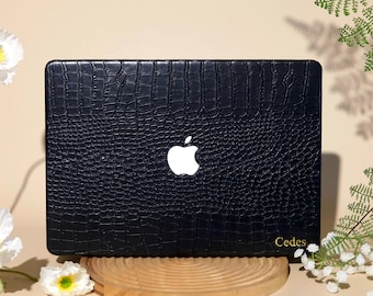 May include: A black laptop cover with a textured crocodile skin pattern and a central white Apple logo. The word Cedes is printed in small gold letters on the bottom right corner. The cover is positioned on a round wooden base.