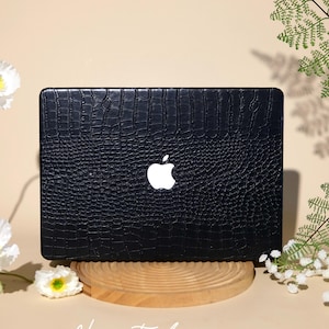 Puede incluir: Una funda para portátil negra con un patrón de textura de piel de cocodrilo y un recorte central que revela el logotipo de Apple plateado. La funda está colocada sobre un soporte de madera circular, con el texto "Ninetydegree." impreso en blanco.