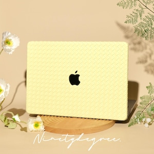May include: A light yellow laptop case with a woven texture and a black Apple logo. The laptop is resting on a wooden stand, with white flowers and greenery surrounding it. The text "Ninetydegree" is written in cursive.