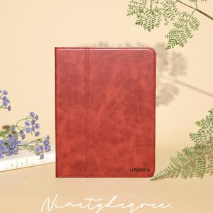 Puede incluir: Una funda rectangular para tableta de piel sintética de color rojo oscuro con un acabado mate liso y una costura vertical en el lado izquierdo. La palabra UNHEA está grabada en la esquina inferior derecha. La funda está colocada sobre una superficie.