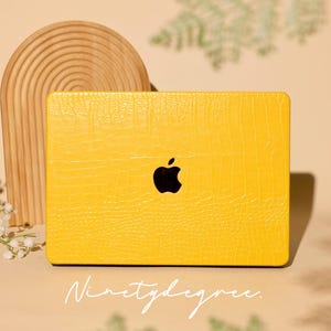 Puede incluir: Una funda amarilla para portátil con un patrón texturizado similar a la piel de cocodrilo. La funda presenta un logotipo de Apple negro y el texto manuscrito "Ninetydegree". El fondo es neutro, con un arco de madera y vegetación.