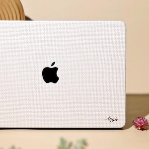 May include: A white laptop skin with a textured, woven-like surface pattern applied to the lid of a laptop. The skin features a black Apple logo in the centre and the name Angie printed in a black script font in the bottom right corner.