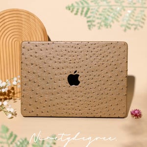 Könnte beinhalten: Eine beigefarbene Laptop-Hartschalenhülle mit einer strukturierten Straußenleder-Optik und einem mittigen Ausschnitt für das Apple-Logo. Die Oberfläche weist ein durchgehendes Punktmuster auf. Der Text Ninetydegree. ist unten zu sehen.