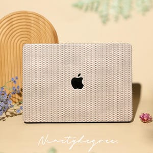 Puede incluir: Una funda para portátil con un patrón de textura de tela tejida en beige y blanco aplicada a la tapa de un portátil cerrado. El logotipo negro de Apple es visible en el centro. El texto Ninetydegree está escrito en cursiva blanca debajo.