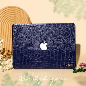 May include: A dark blue laptop cover with a textured crocodile skin pattern. The cover features a central white Apple logo cutout and the name Cedes printed in small gold letters in the bottom right corner. The surface has a glossy finish.