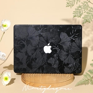 Könnte beinhalten: Schwarze Laptop-Hülle mit Blättermuster und weißem Apple-Logo. Die Hülle wird auf einem Holzständer mit dem kursiven Text "Ninetydegree" präsentiert. Der Hintergrund umfasst weiße Blumen und grünes Laub.