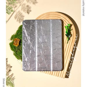 Peut inclure: Étui pour iPad en acrylique argenté. L'étui a un aspect métallique texturé. Les mots "Acrylic iPad case", "Silver color", "High quality material" et "Perfectly fits your device" sont visibles.
