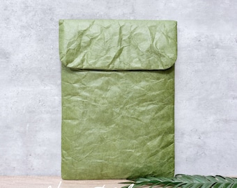 May include: A green, crinkled paper laptop sleeve with a fold-over flap. The sleeve is sitting on a wooden surface with a green leaf from a plant.