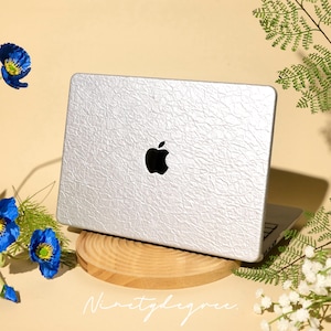 May include: Silver laptop with a textured surface and a black Apple logo, resting on a wooden stand. The laptop is open, revealing the keyboard. The background is a light beige color, with blue flowers and greenery.