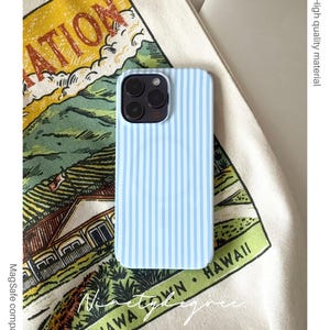 Könnte beinhalten: Hellblaue iPhone-Hülle mit vertikalen weißen Streifen. Die Hülle wird auf einem gemusterten Stoff mit dem Text "WAHIAWA TOWN. HAWAII" gezeigt. Die Hülle ist MagSafe-kompatibel und aus hochwertigem Material gefertigt.
