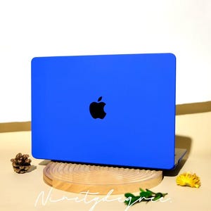 Può includere: Un laptop blu con il logo Apple nero è esposto su un supporto di legno. Una pigna e un fiore giallo sono posti su entrambi i lati del laptop. Il testo "Ninetydegree" è scritto in corsivo sotto.