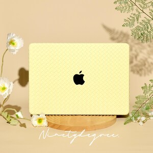 May include: A pale yellow laptop skin featuring a textured, woven-look pattern and a centred black Apple logo. The rectangular skin is displayed on a circular wooden stand, with the word "Ninetydegree." printed in white script below the laptop.