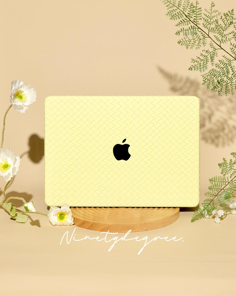 May include: A pale yellow laptop skin featuring a textured, woven-look pattern and a centred black Apple logo. The rectangular skin is displayed on a circular wooden stand, with the word "Ninetydegree." printed in white script below the laptop.