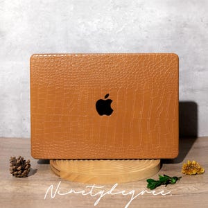 May include: A brown laptop case with a crocodile skin texture and a black Apple logo. The case is displayed on a wooden stand with the word "Ninetydegree" in white script.