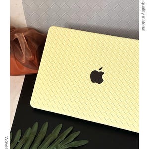 May include: A pale yellow laptop cover featuring a woven texture pattern. The cover has a cutout in the centre revealing a black Apple logo. The text on the image includes Chic colour, High quality material, Woven style, and Perfectly fits your device.