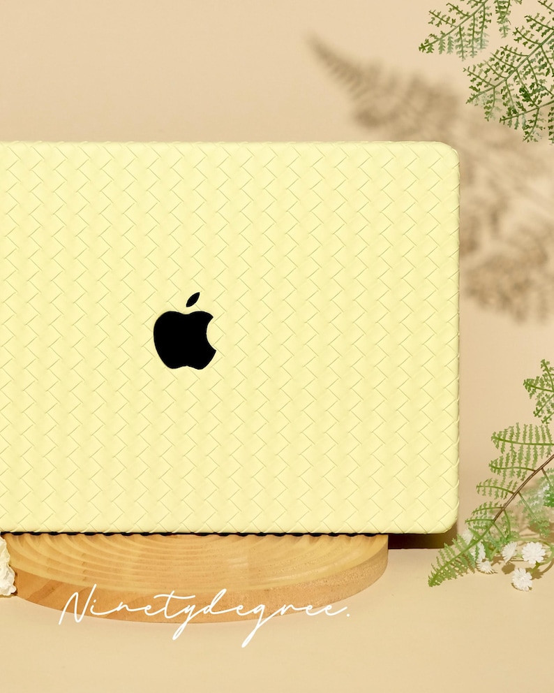 May include: A pale yellow laptop cover featuring a woven, basket-weave texture across the entire surface. A black Apple logo is centred on the cover. The text "Ninetydegree." is printed in a white, cursive script at the bottom of the cover.
