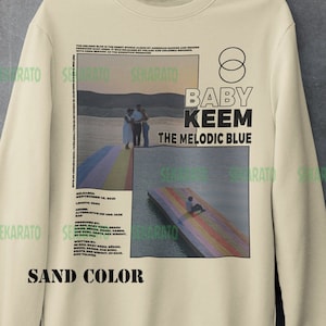 May include: A beige sweatshirt with a graphic print of a sunset scene and the text "BABY KEEM THE MELODIC BLUE" in black. The graphic also includes a rainbow-colored walkway and a person sitting on a floating platform. The text "SAND COLOR" is printed below the graphic.