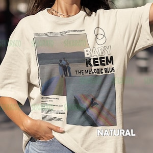 May include: A beige t-shirt with a graphic print of a photo of two people standing on a beach with a rainbow in the background. The text "BABY KEEM THE MELODIC BLUE" is printed on the shirt. The word "NATURAL" is printed on the bottom of the shirt.