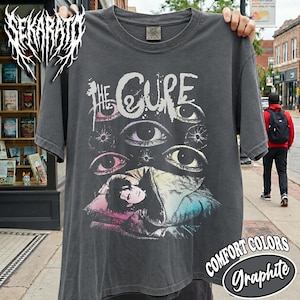 May include: A charcoal gray short-sleeve t-shirt featuring a graphic print of the band name The Cure in white lettering above multiple stylized eyes and an illustration of a person resting on a pillow with pink, blue, and white color accents.