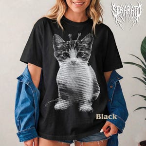 Op de afbeelding: Zwart T-shirt met een halftone afbeelding van een kat met antennes. Het woord "Black" staat onderaan gedrukt. Onderdeel van een kledingcollectie.
