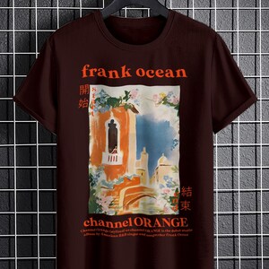 May include: A brown t-shirt with a graphic print of a painting of a building with a balcony and flowers. The text "frank ocean" is printed in orange above the painting. The text "channel ORANGE" is printed in orange below the painting. The text "開始" and "結束" are printed in orange on either side of the painting.