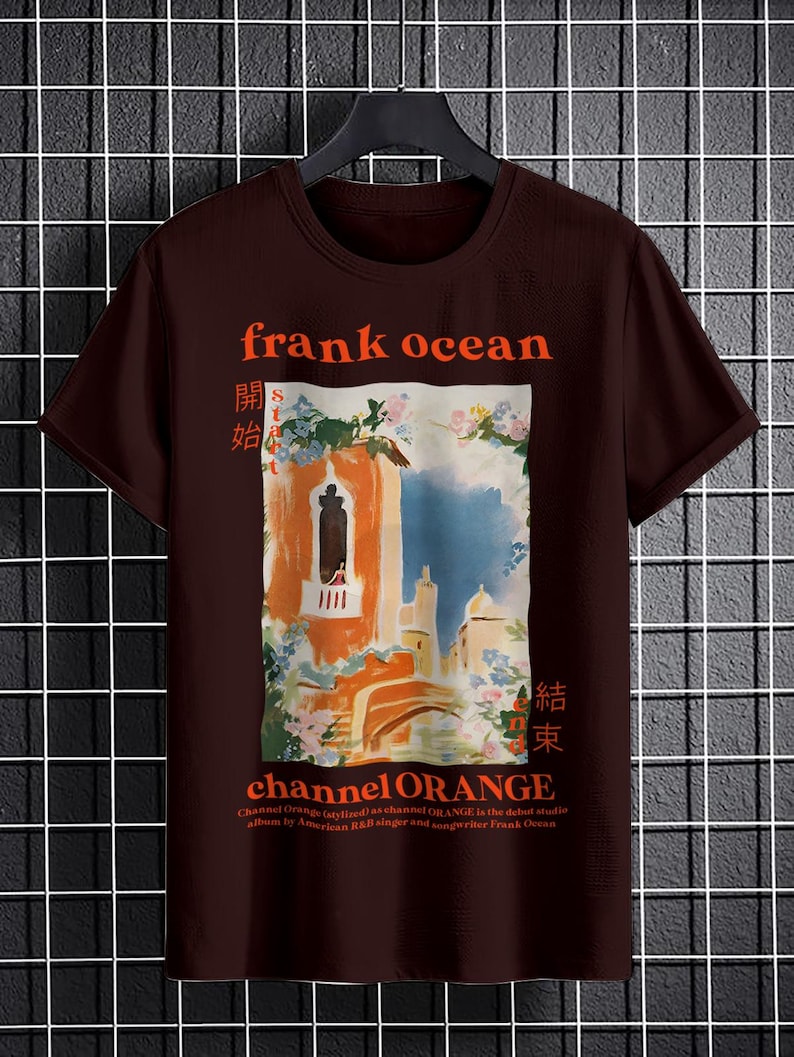 May include: A brown t-shirt with a graphic print of a painting of a building with a balcony and flowers. The text "frank ocean" is printed in orange above the painting. The text "channel ORANGE" is printed in orange below the painting. The text "開始" and "結束" are printed in orange on either side of the painting.