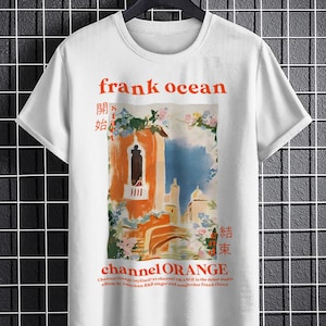 May include: White t-shirt with a graphic print of a painting of a building with a blue sky and flowers. The text "frank ocean" is printed in orange above the painting. The text "channel ORANGE" is printed in orange below the painting. The text "開始" and "結束" are printed in black on either side of the painting.