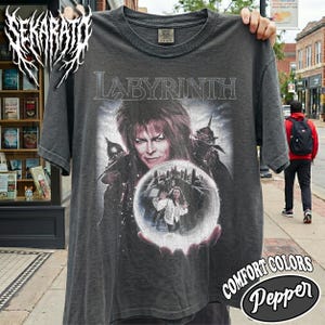 May include: A charcoal grey short-sleeve t-shirt featuring a graphic print from the film Labyrinth. The design displays a large image of Jareth the Goblin King holding a crystal ball with Sarah inside, with the word Labyrinth printed above.