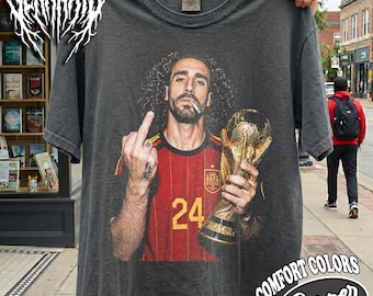 Może przedstawiać: Ciemnoszary t-shirt z krótkim rękawem z nadrukiem graficznym przedstawiającym piłkarza z długimi kręconymi włosami, brodą i papierosem w ustach. Zawodnik ma na sobie czerwoną koszulkę z numerem 24 i trzyma złote trofeum, wykonując gest.