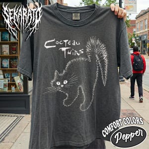 May include: A charcoal gray short-sleeve t-shirt featuring a white graphic print of a stylized, arched-back cat with long whiskers and a fluffy tail. The text "Cocteau Twins" is printed in a distressed white font above the cat illustration.