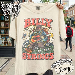 Może przedstawiać: Beżowy t-shirt z grafiką przedstawiającą osobę w kapeluszu i z plecakiem, grającą na gitarze akustycznej. Wzór zawiera góry, kwiaty, motyla oraz czerwony napis BILLY STRINGS. Koszulka posiada okrągły dekolt i krótkie rękawy.