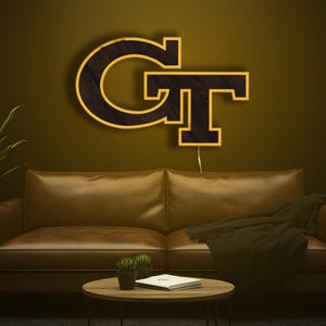 May include: A lit wooden sign in the shape of the letters "GT" in black with a gold outline, mounted on a wall. The sign is above a brown leather couch with a black blanket. A small round table with plants and a book is in front of the couch.