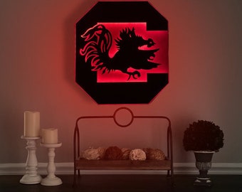 May include: A black and red illuminated wall decor featuring a rooster silhouette inside a large letter "C". The decor is mounted on a wall with two white candle holders and a small wooden shelf with decorative balls.