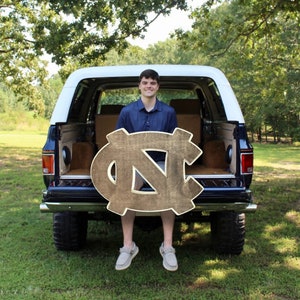 Puede incluir: Una persona joven sentada en la parte trasera de una camioneta azul sosteniendo un letrero de madera grande con las letras "NC" pintadas en oro.