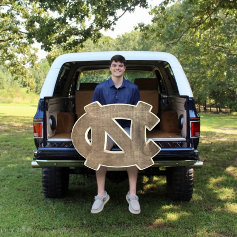 Puede incluir: Una persona joven sentada en la parte trasera de una camioneta azul sosteniendo un letrero de madera grande con las letras "NC" pintadas en oro.