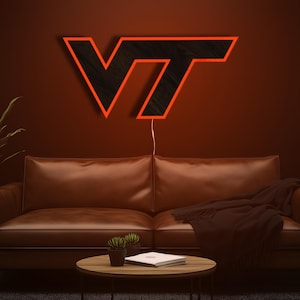 May include: A lit wooden sign in the shape of the letters "VT" with a black interior and orange outline. The sign is mounted on a dark brown wall above a brown leather couch. A small round table and plants are in front of the couch.