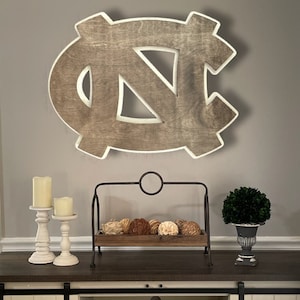 Puede incluir: Una decoraci&oacute;n de pared de madera con la forma del logotipo de los Tar Heels de la Universidad de Carolina del Norte. El logotipo est&aacute; pintado en color marr&oacute;n claro con un contorno blanco. El logotipo est&aacute; colgado en una pared gris sobre una mesa de madera con dos candelabros blancos y un soporte met&aacute;lico decorativo con una bandeja de madera.