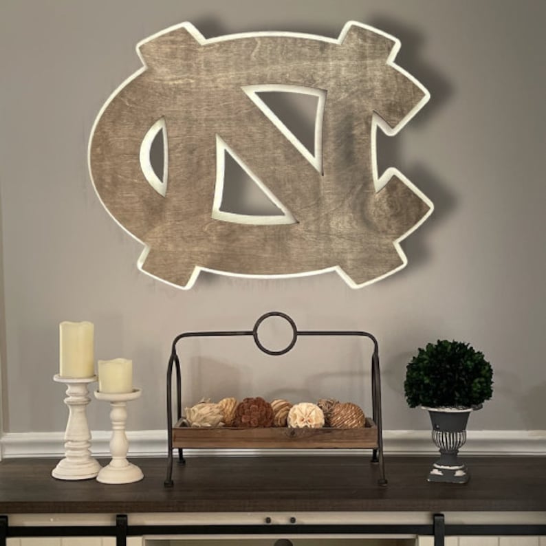 Puede incluir: Una decoraci&oacute;n de pared de madera con la forma del logotipo de los Tar Heels de la Universidad de Carolina del Norte. El logotipo est&aacute; pintado en color marr&oacute;n claro con un contorno blanco. El logotipo est&aacute; colgado en una pared gris sobre una mesa de madera con dos candelabros blancos y un soporte met&aacute;lico decorativo con una bandeja de madera.