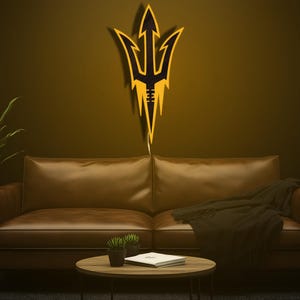 May include: A gold and black trident-shaped wall decor is mounted on a dark wall above a brown leather sofa. The trident has a central arrow shape with three prongs and a football-like design at the bottom. A round coffee table and plants are in front of the sofa.