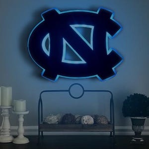 Puede incluir: Una decoraci&oacute;n de pared iluminada azul y negra con el logotipo de los Tar Heels de la Universidad de Carolina del Norte. El logotipo es una "N" y una "C" estilizadas entrelazadas. La decoraci&oacute;n est&aacute; iluminada desde atr&aacute;s con una luz azul.