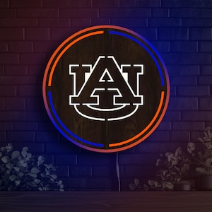 May include: A round wooden sign featuring the Auburn University logo in white. The sign is encircled by a blue and orange neon light, set against a dark brick wall. The logo is the letters "AU" in a stylized font.