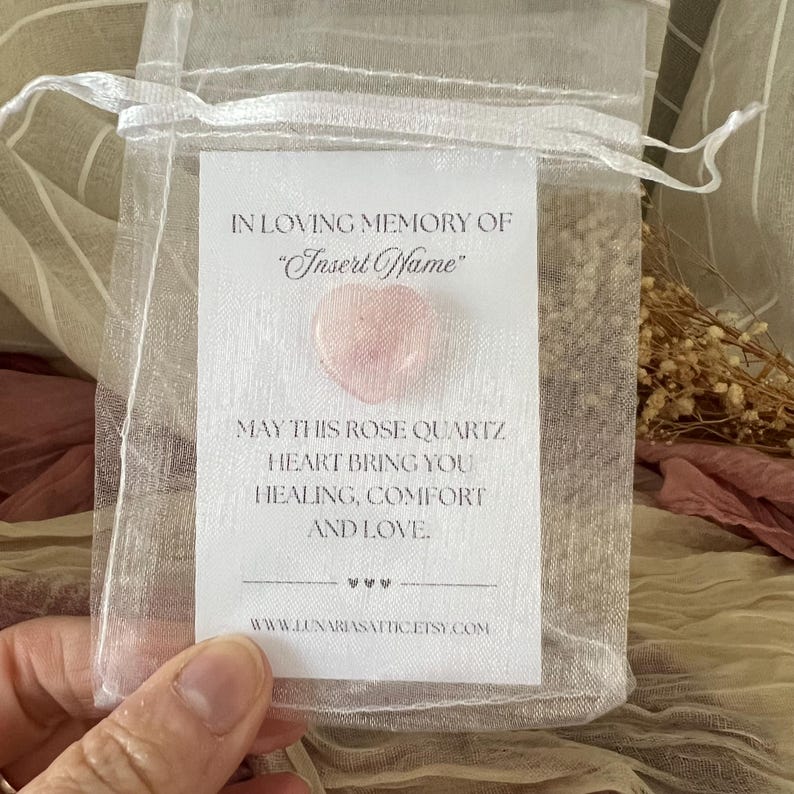 Op de afbeelding: Een kleine, transparante organzazak met een roze rozenkwartshart en een witte kaart. De kaart luidt "TER LIEFDEVOLLE HERINNERING AAN 'Insert Name'" en "MOGE DIT ROZENKWARTS HART JE GENEZING, TROOST EN LIEFDE BRENGEN."