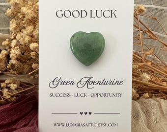 Op de afbeelding: Een hartvormige groene aventurijnsteen ligt op een witte kaart. De kaart toont bovenaan "GOOD LUCK", met daaronder "Green Aventurine" en "SUCCESS - LUCK - OPPORTUNITY". De Etsy shop URL staat onderaan.