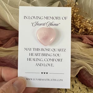 Op de afbeelding: Een lichtroze rozenkwarts hartvormige steen op een witte kaart. De kaart leest "In Loving Memory of 'Insert Name'" en "May this rose quartz heart bring you healing, comfort, and love." De Etsy shop URL is ook zichtbaar.