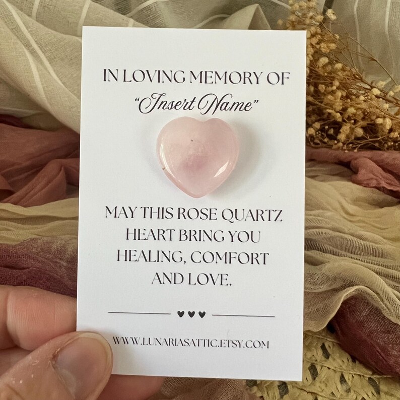 Op de afbeelding: Een lichtroze rozenkwarts hartvormige steen op een witte kaart. De kaart leest "In Loving Memory of 'Insert Name'" en "May this rose quartz heart bring you healing, comfort, and love." De Etsy shop URL is ook zichtbaar.