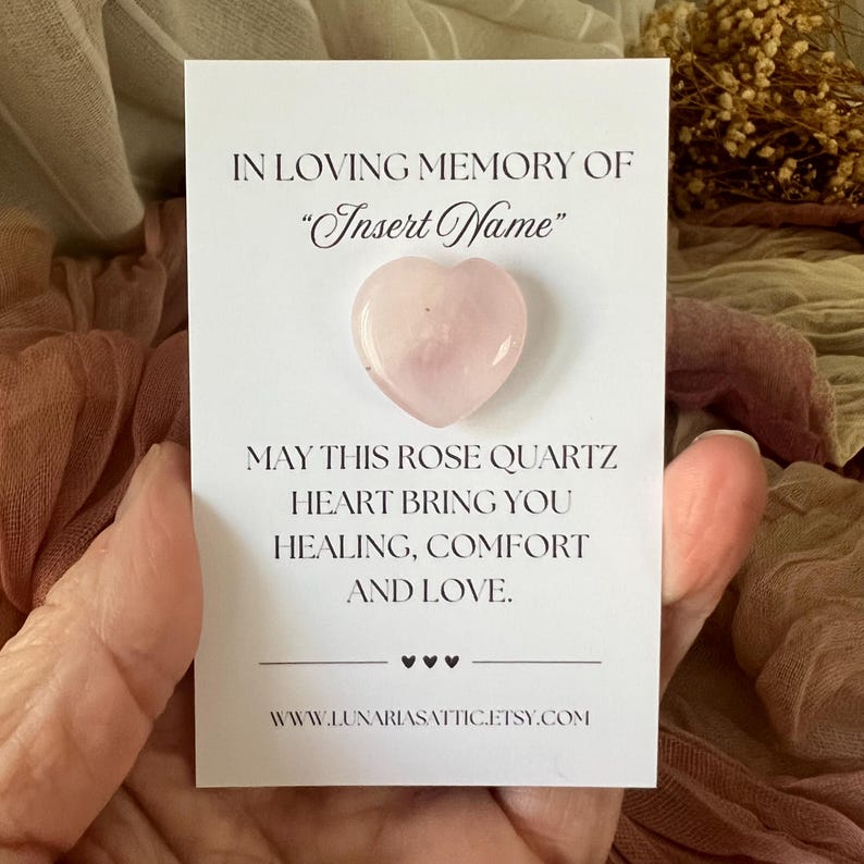 Op de afbeelding: Een rozenkwarts hartvormige steen op een witte kaart. De kaart leest "In Loving Memory Of" en "Insert Name". Hieronder staat "May this rose quartz heart bring you healing, comfort, and love."