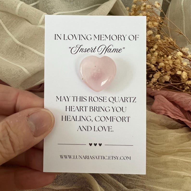 Op de afbeelding: Een lichtroze rozenkwarts hartvormige steen op een witte kaart. De kaart leest "IN LOVING MEMORY OF 'Insert Name'" en "MAY THIS ROSE QUARTZ HEART BRING YOU HEALING, COMFORT AND LOVE."