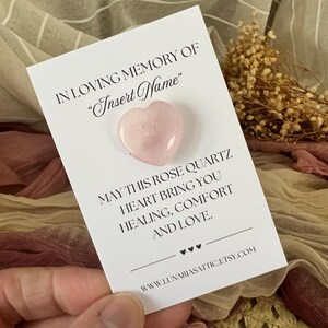 Op de afbeelding: Een roze hartvormige rozenkwartssteen ligt op een witte kaart. De kaart leest "IN LOVING MEMORY OF 'Insert Name'" en "MAY THIS ROSE QUARTZ HEART BRING YOU HEALING, COMFORT AND LOVE." Het websiteadres is ook zichtbaar.